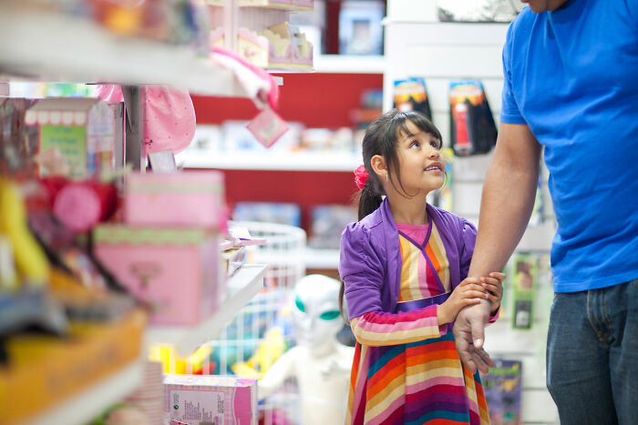 Young girl holding adult's hand in a toy store, surrounded by colorful items and a visible alien figure in the background.