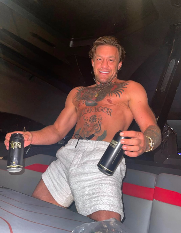 Shirtless man with tattoos smiling and holding two beverage cans in a dimly lit indoor setting.