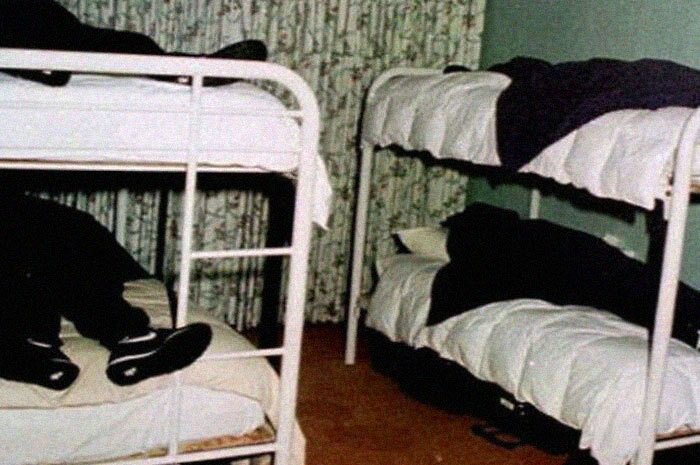 Dimly lit room with white metal bunk beds, dark-clothed figures lying still, evoking terrifying and horrific backstories. - 39