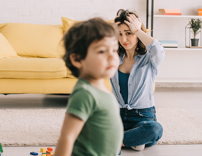Woman sitting on floor stressed while babysitting toddler in living room with yellow couch in background - 1