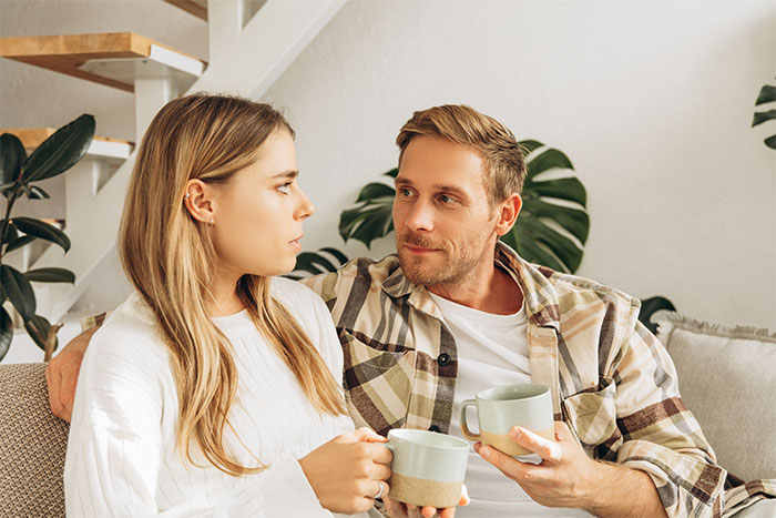 Woman questions marriage while holding coffee cup, looking concerned as husband gazes at her thoughtfully on couch. Woman questions marriage while holding coffee cup, looking concerned as husband gazes at her thoughtfully on couch.