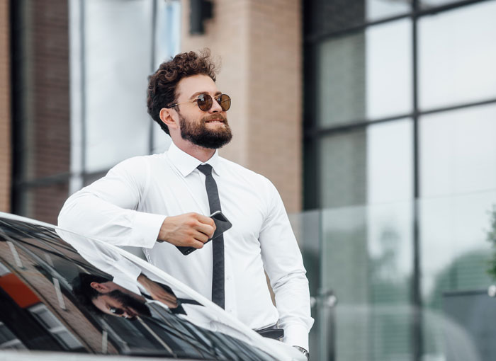 Man in a white shirt and black tie holding car keys, standing by luxury car, illustrating unexpected things about the rich.