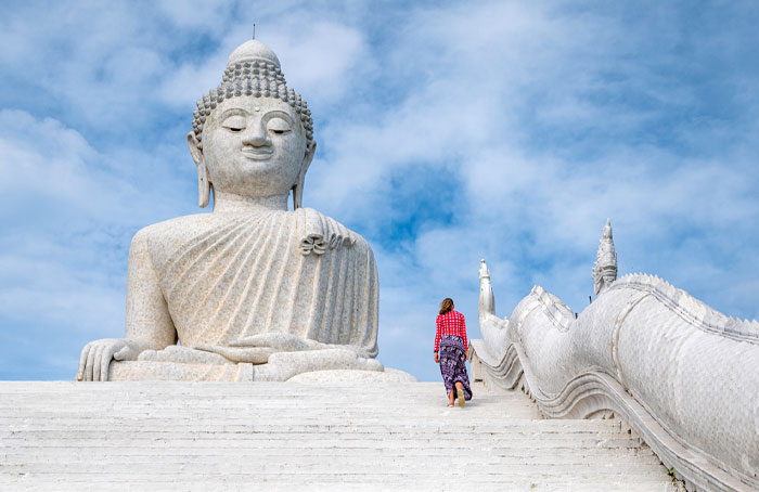 Woman walking up stairs beside a large Buddha statue under a clear blue sky in a scenic outdoor setting. - 6
