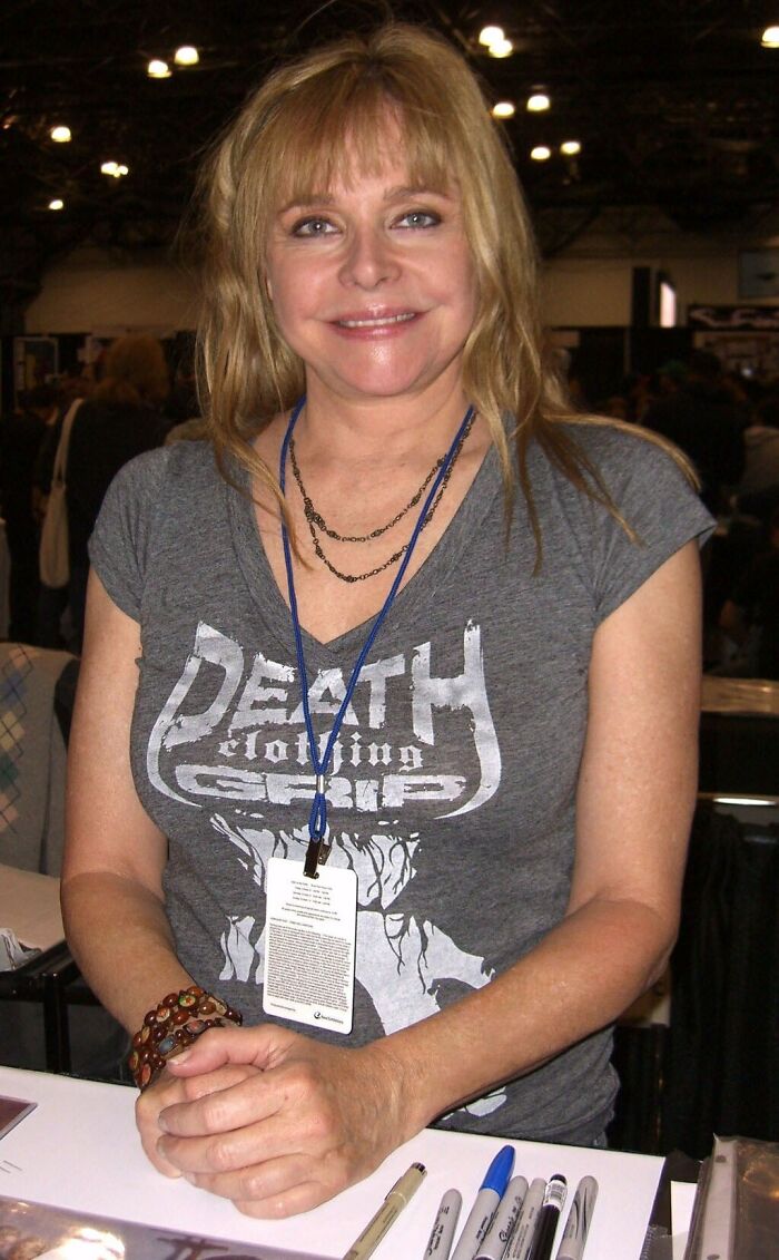 Bond Girl at 70 smiling and seated at a table with pens, wearing a gray graphic t-shirt and layered necklaces. Bond Girl at 70 smiling and seated at a table with pens, wearing a gray graphic t-shirt and layered necklaces.