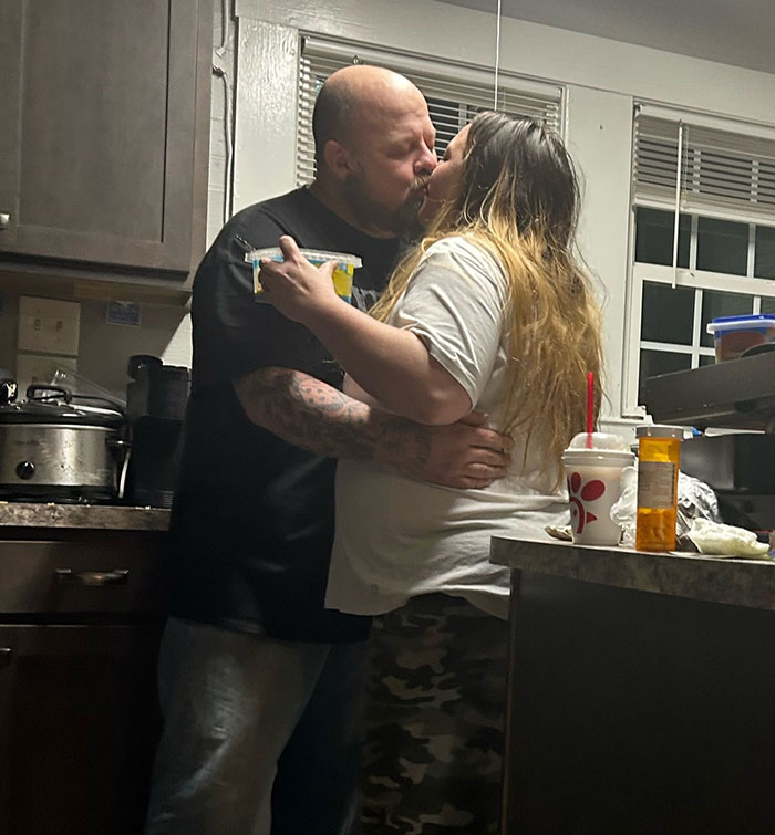 Couple embracing and kissing in a kitchen, with kitchen appliances and food containers visible on counters.