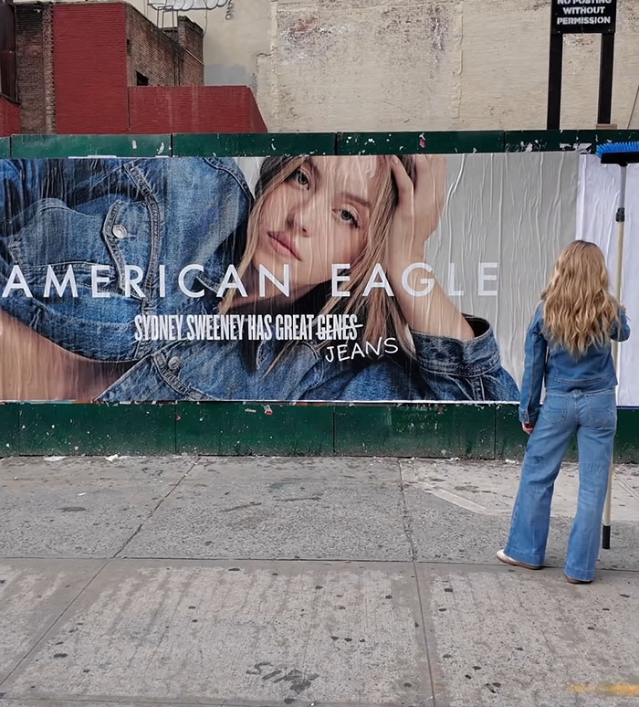 Woman in denim jeans and jacket looking at a large American Eagle jeans ad featuring Sydney Sweeney outdoors. - 12