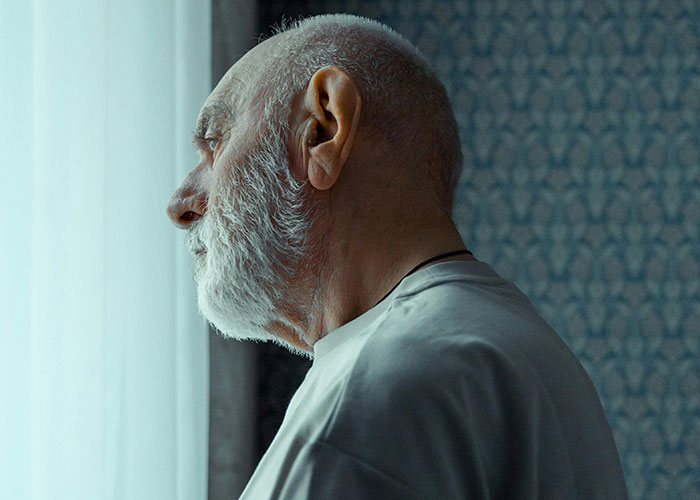 Elderly man with a white beard looking pensively out a window in a dimly lit room reflecting on a difficult relationship.