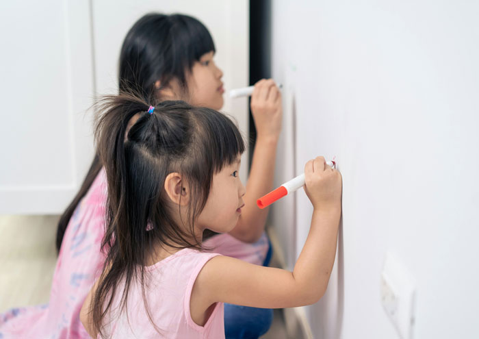 Two children drawing on a white wall with markers, showcasing playful home activities beyond oldschool home trends.
