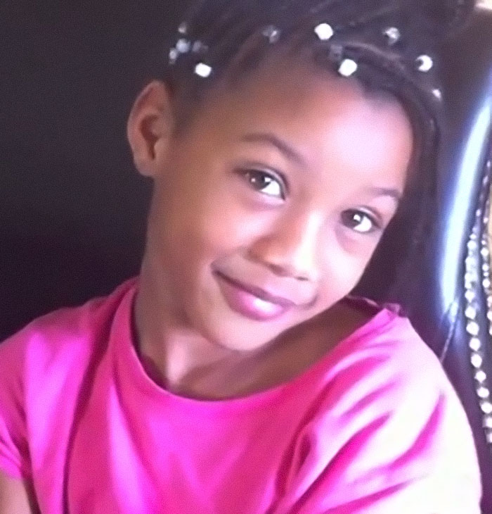 Young girl with braided hair and beads, smiling softly while wearing a bright pink shirt against a dark background