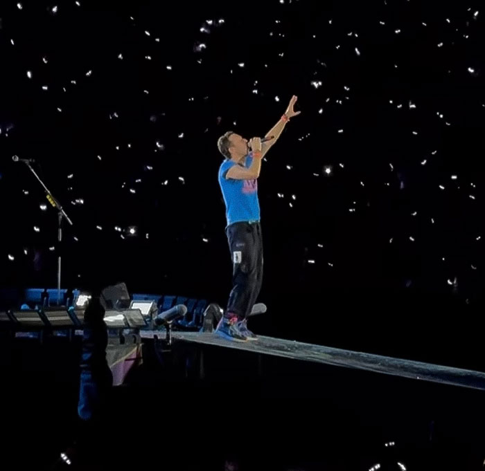 Coldplay lead singer performing on stage with lights from audience phones creating a starry effect during concert.