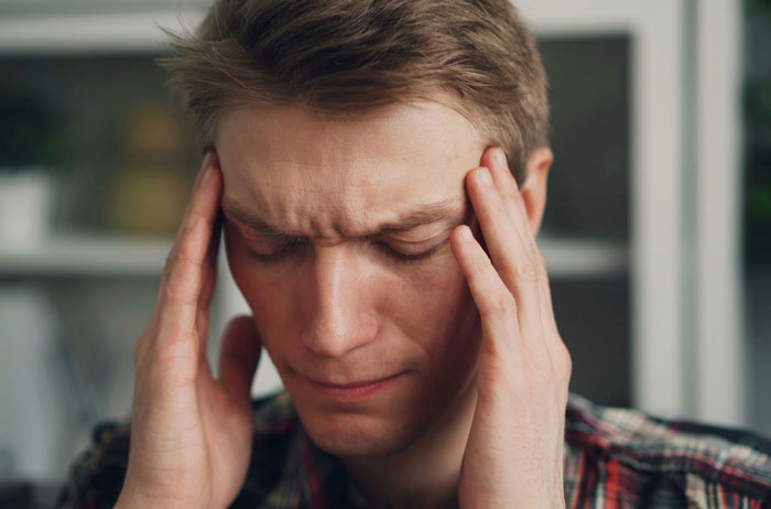 Young man with closed eyes holding temples, appearing stressed and upset in a home setting.