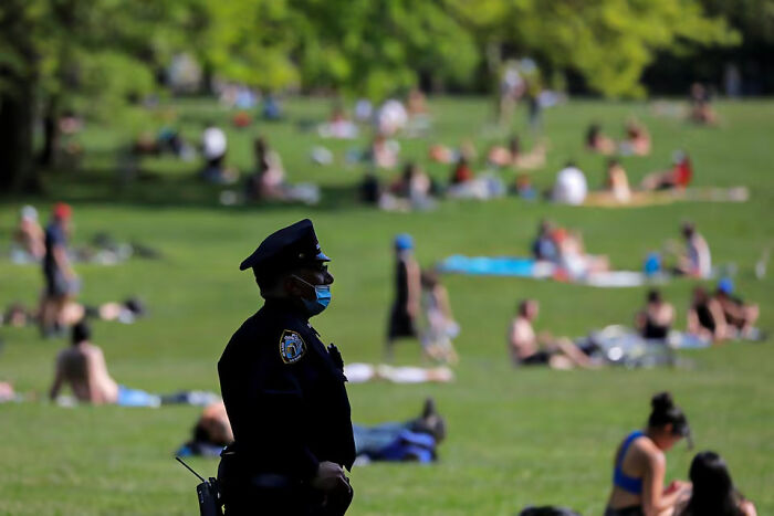 Police officer wearing a mask standing in a park with people relaxing in the background representing job consequences.