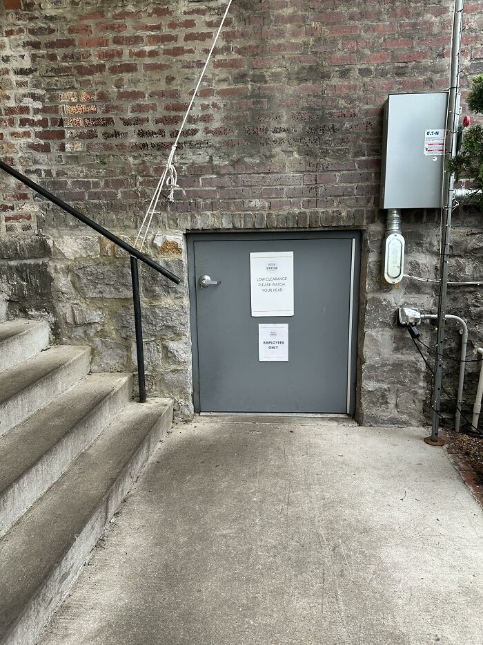 Small gray door with low clearance sign under brick wall near concrete stairs showing failed home creativity attempt