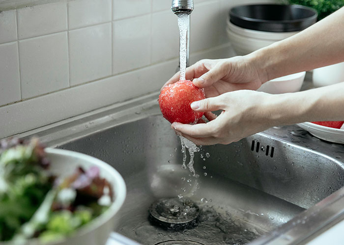 Person washing a tomato under running water in a kitchen sink showing normal family habits and routines.