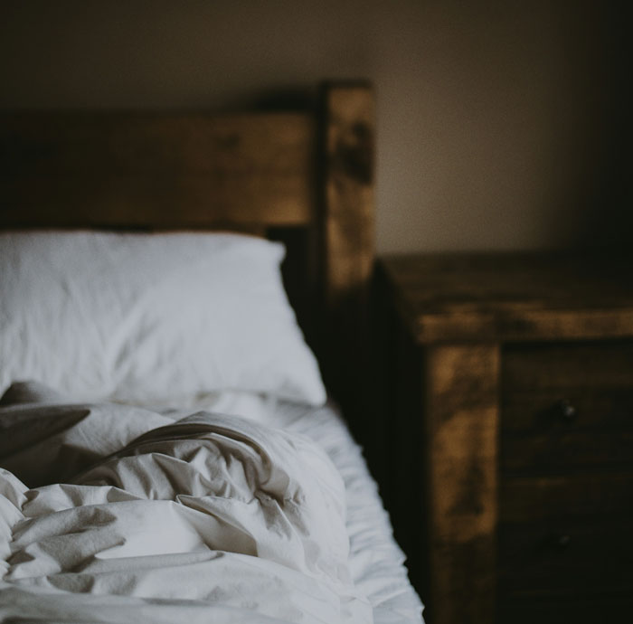 Unmade bed with white sheets and a rustic wooden headboard and nightstand in dimly lit room evoking real-life horror stories - 15