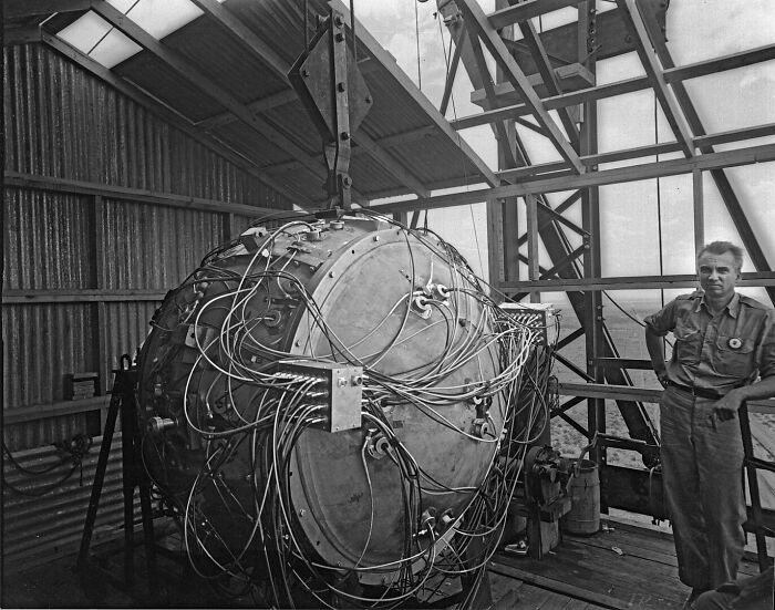 Black and white photo of a man standing next to a large experimental device with many wires inside an industrial building, illustrating interesting rumors turned true.