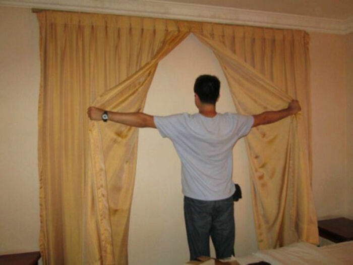 Person standing in a bedroom holding curtains open to reveal a blank wall, a creative home design fail.