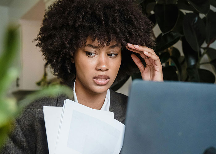 Young professional woman looking stressed at laptop, holding papers, illustrating micromanagement in the workplace.