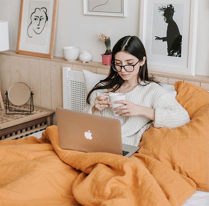 Young woman in glasses using laptop in bed with orange blanket, reflecting on not sharing important news with dad.