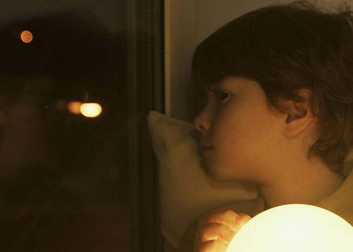 Child holding a glowing ball, looking out a window at night, reflecting on bizarre family behaviors considered normal.