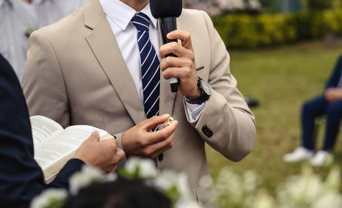 Man in beige suit holding microphone and wedding ring during outdoor ceremony with officiant holding a book. - 32