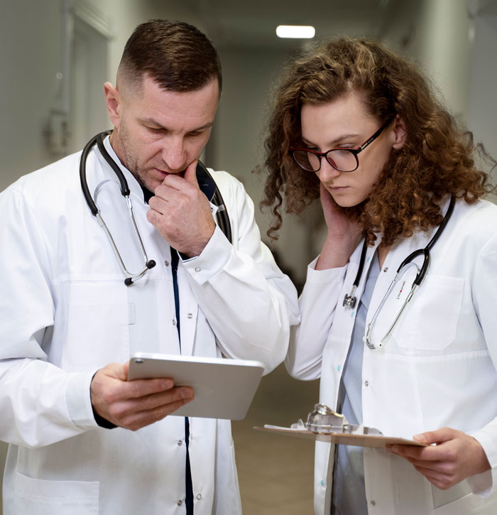 Two doctors in white coats reviewing patient information on a tablet and clipboard in a hospital hallway. - 35