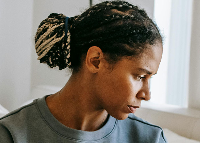 Young woman with braided hair looking down pensively by a window, reflecting on worst cheating stories and emotional pain.
