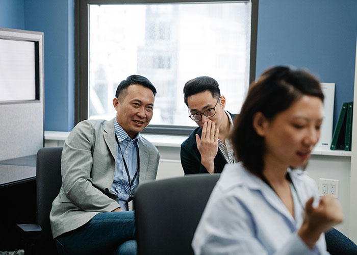 Two men whispering and watching a woman working, illustrating micromanagement signs in a tense workplace environment.