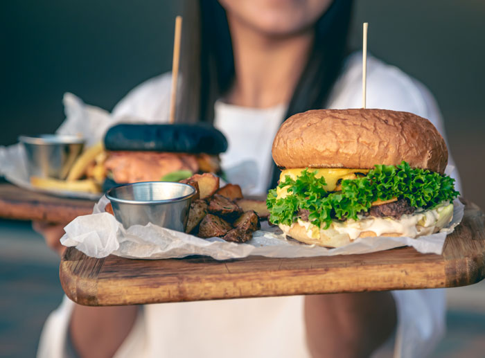 Woman holding wooden boards with burgers and side dishes in a casual dining setting, related to public spaces comments topic.