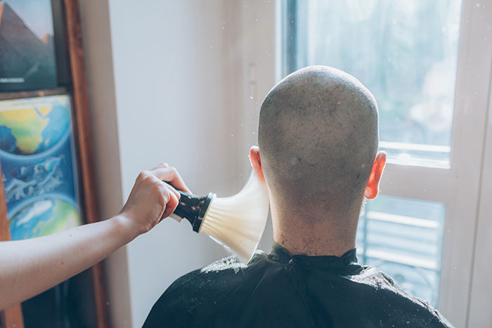 Teen boy with shaved head being brushed after haircut in support of family member with cancer at home near window.