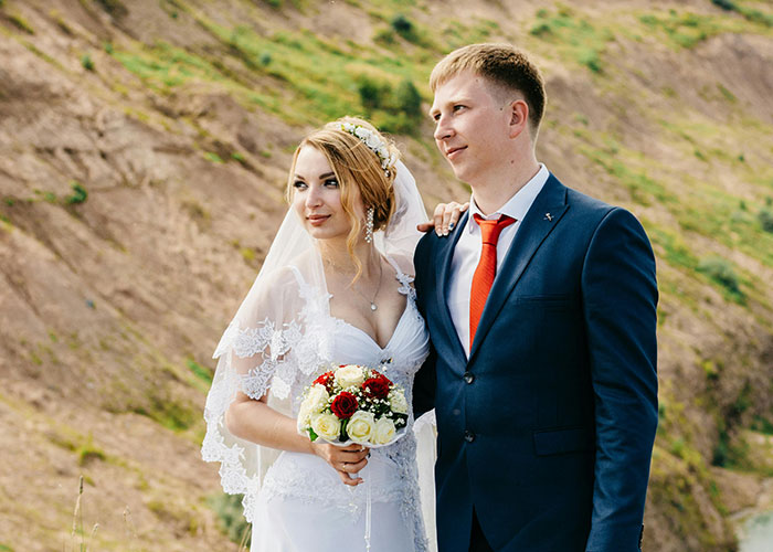 Bride in a white lace dress and groom in a blue suit with red tie standing outdoors on their wedding day. - 6