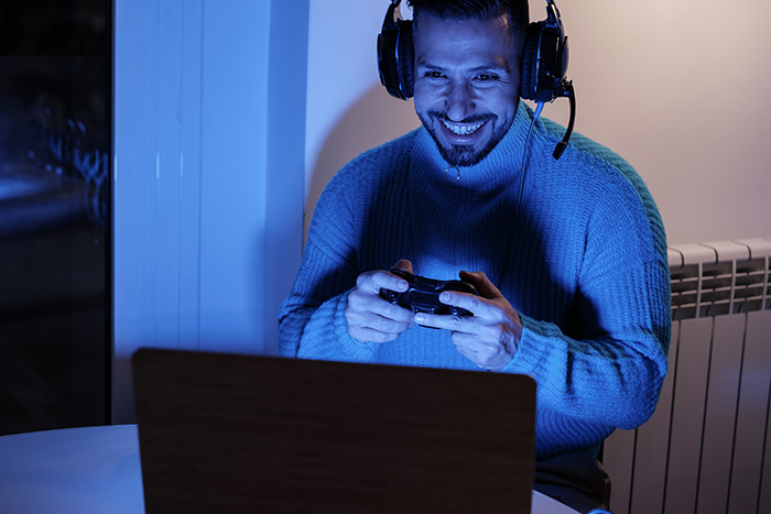 Man wearing headphones and holding a game controller, smiling while playing video games at night on a laptop. - 12