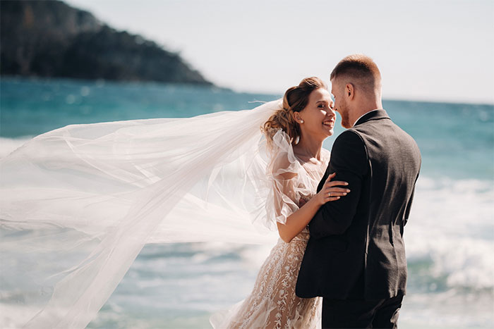 Bride and groom embracing on beach with bride’s veil flowing, highlighting wedding aesthetic under 102°F heat conditions. Bride and groom embracing on beach with bride’s veil flowing, highlighting wedding aesthetic under 102°F heat conditions.
