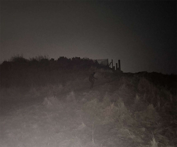 Dark night scene showing a mysterious figure in a full-body cat suit crawling on a grassy hill near a fence.