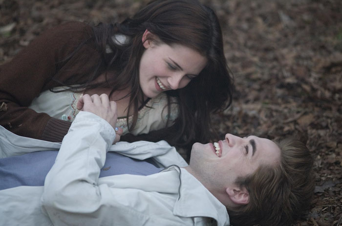 Robert Pattinson and Kristen Stewart in a Twilight intimate scene, smiling closely while lying on the forest floor.