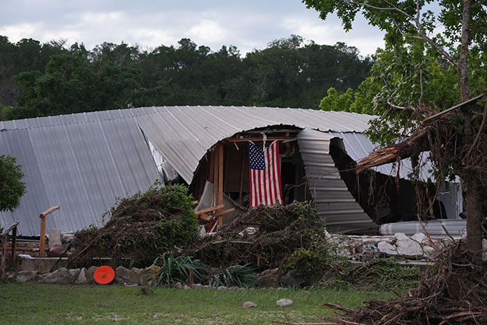 Damaged camp building with American flag visible, surrounded by fallen trees and debris after natural disaster at Camp Mystic.