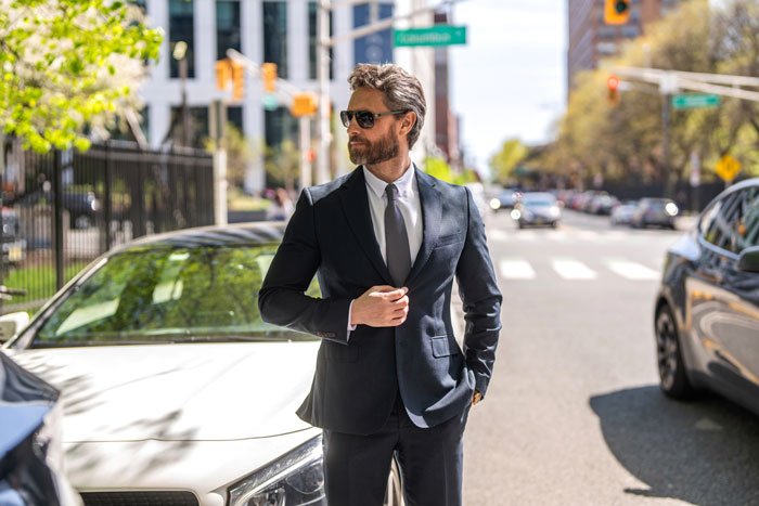 Man in a dark suit and sunglasses standing confidently near luxury cars in a city, symbolizing wealthy lifestyle and marriage.
