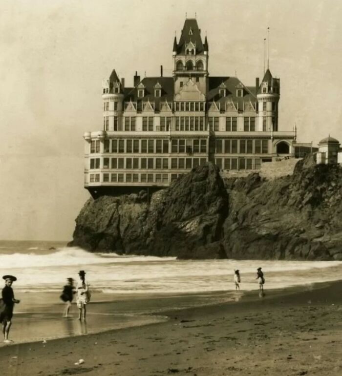 Old architecture featuring a grand historic building perched on a rocky coastline with people walking on the beach.