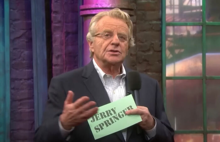 Man holding a card with Jerry Springer text, hosting a talk show about unhinged family lore stories.