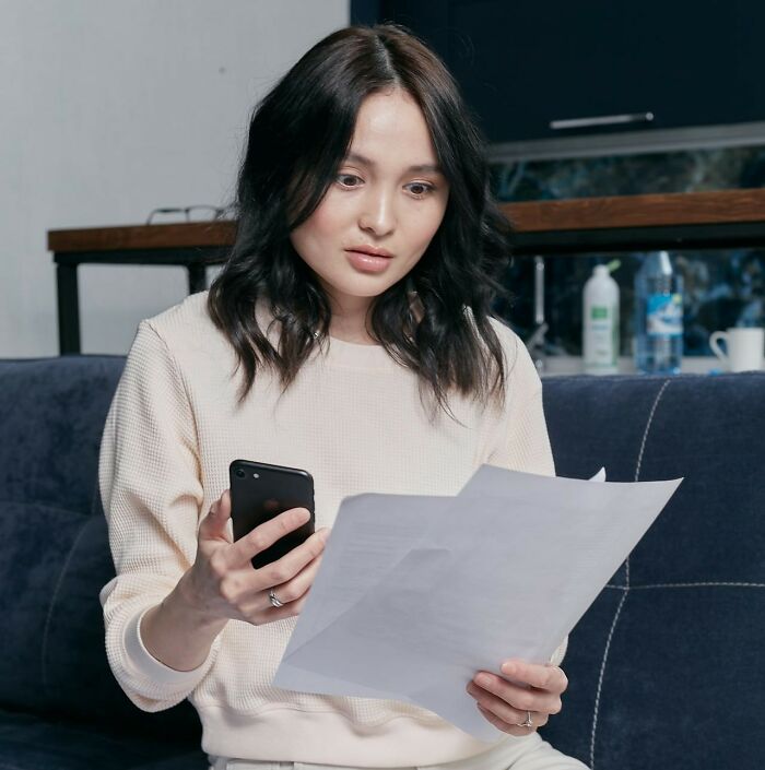 Young woman looking surprised while reading a printed paper and holding a smartphone, reflecting messed up school incidents emotions. - 25