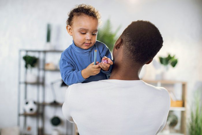 Mom holding toddler while babysitting at home, highlighting conflict over teen asking to be paid for babysitting.