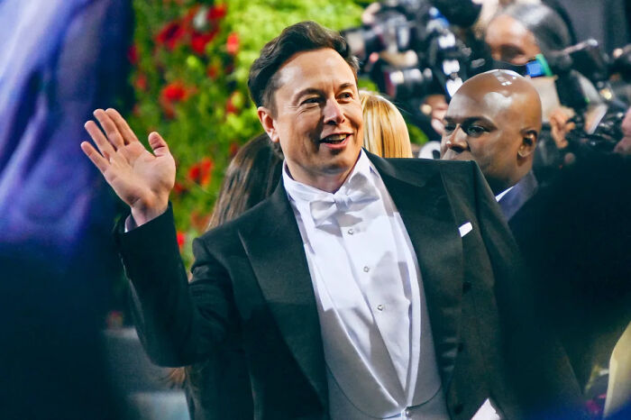 Elon Musk in a tuxedo waving at an event, surrounded by people and photographers, illustrating consequences of stupidity and job loss.