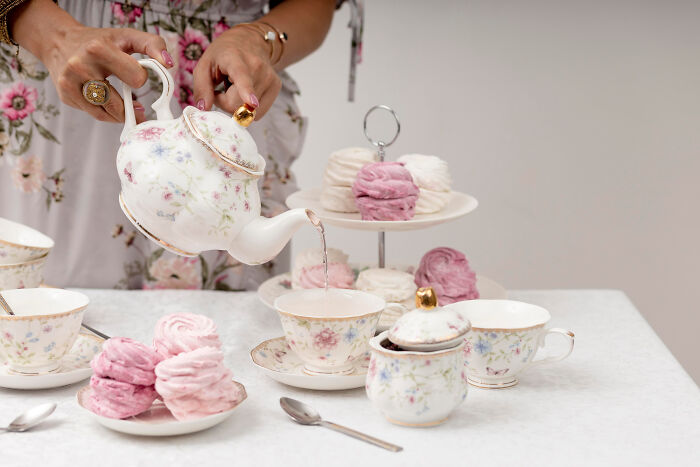 Person pouring tea from a vintage floral teapot into matching teacups during a nostalgic tea time with pastel desserts.