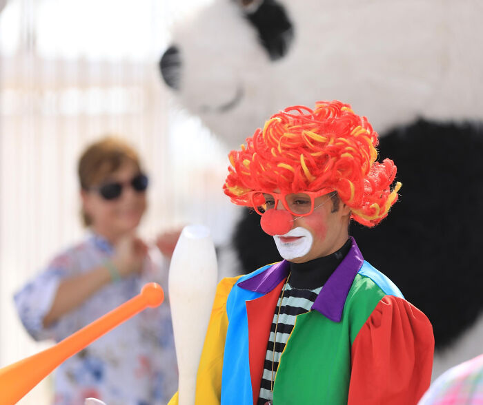Clown with bright orange wig and red nose performing in colorful outfit at an event with people in the background. - 2
