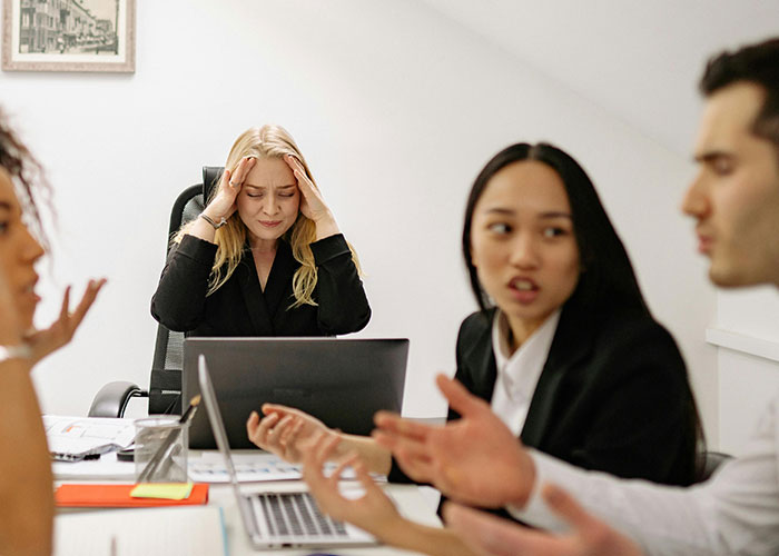 Stressed woman at desk experiencing micromanagement issues while colleagues argue in a tense workplace meeting environment.