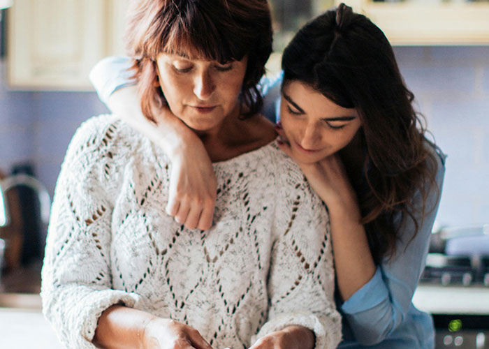 Two women in a kitchen sharing a close moment, reflecting on bizarre family habits considered normal.