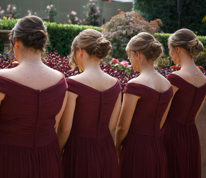 Four bridesmaids in matching burgundy dresses with elegant updos standing outdoors in a garden setting. - 54