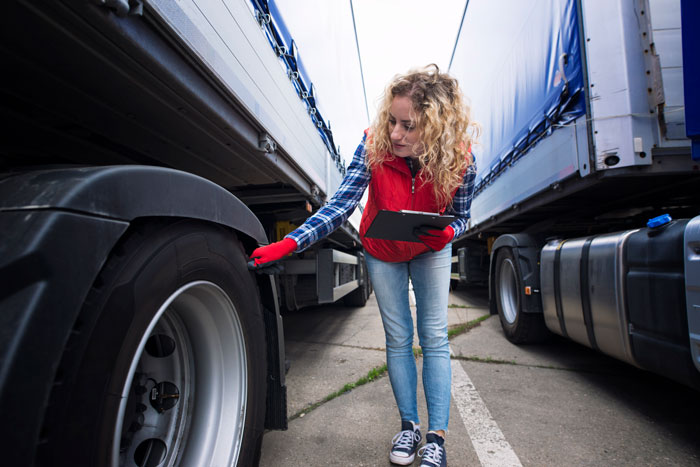 Woman inspecting truck tire between two large trucks in a public space, highlighting women and inappropriate comments from men.