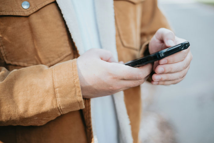 Person wearing brown jacket holding and looking at phone, symbolizing woman leaving to find herself on a dream trip alone.