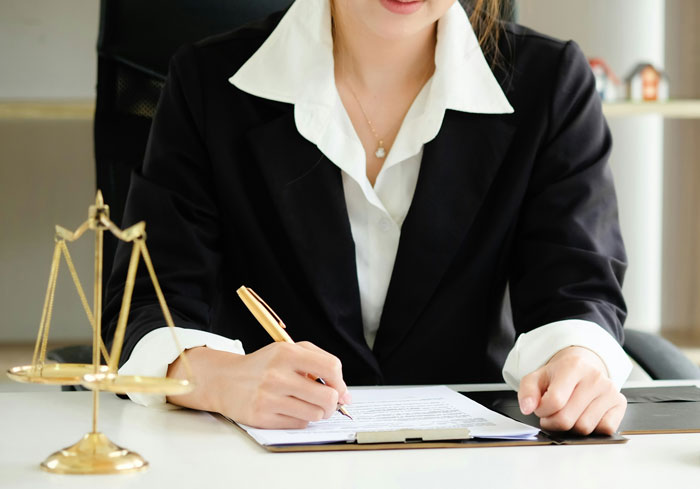 Woman in formal attire signing documents at desk with golden scales of justice nearby, focused and determined.